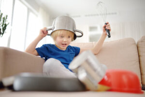 A young child sits on a couch wearing a pot as a hat, holding a whisk. The child looks excited while looking at various pots and pans scattered on the floor, creating a playful atmosphere in a bright living room.