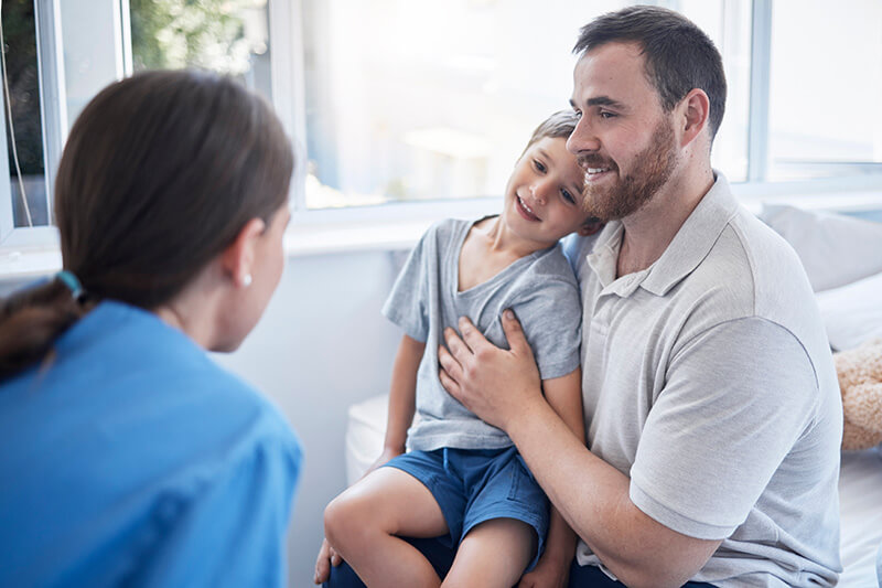 A man smiling while holding a young boy on his lap. They are sitting indoors and facing a woman wearing a blue shirt, who appears to be speaking to them. The room is well-lit, with a window in the background.