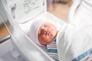 A newborn baby sleeps peacefully in a hospital bassinet. The baby is wrapped in a white blanket and wearing a pink and white striped hat. A small card with footprints and writing is visible in the background.