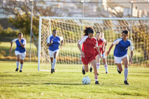 A group of young women playing soccer on a sunny day. One player in a red uniform dribbles the ball towards the goal, while players in blue uniforms run to defend. The goal and field are visible in the background.