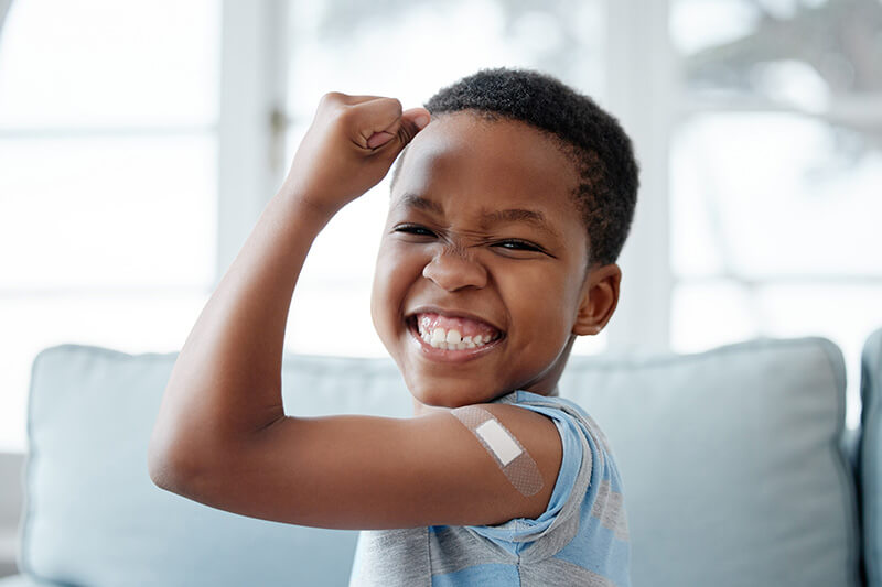 A smiling child flexes their arm while displaying a bandage on their upper arm, suggesting they received a vaccine or shot. The child is indoors, in front of a blurred background.