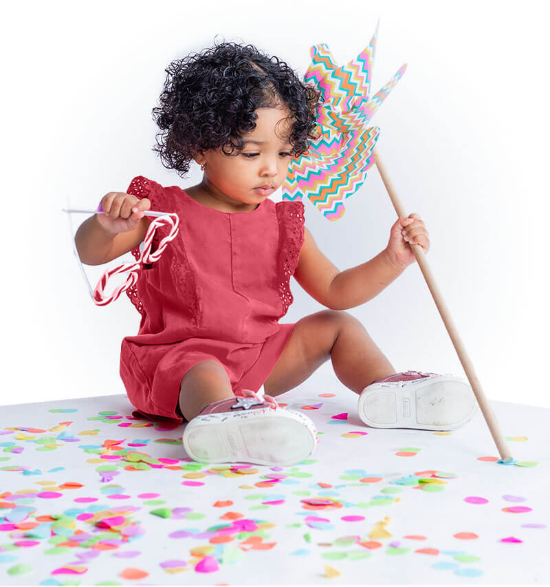 A toddler in a red romper sits on the floor surrounded by colorful confetti. They hold a vibrant pinwheel and a small toy. The background is white, creating a playful and cheerful atmosphere.