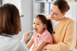 A doctor examines a young girls throat using a tongue depressor. The girl is sitting on a woman's lap, who is wearing a beige sweater. The setting appears to be a medical office.