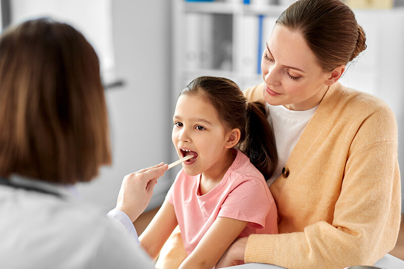 A doctor examines a young girls throat using a tongue depressor. The girl is sitting on a womans lap, who is wearing a beige sweater. The setting appears to be a medical office.