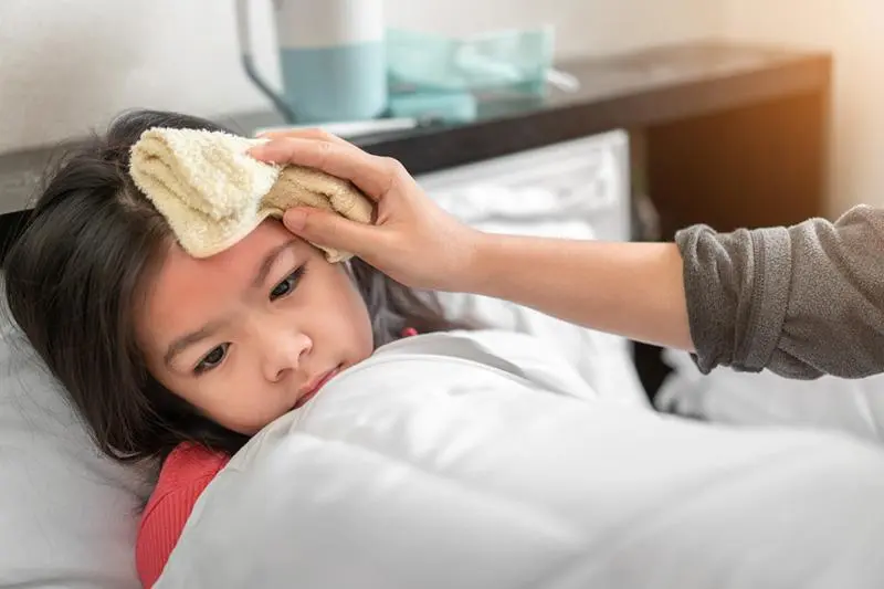 A child lies in bed with a concerned expression, wrapped in a white blanket. A hand is placing a damp cloth on the childs forehead. A bedside table with a tissue box and a kettle is in the background.